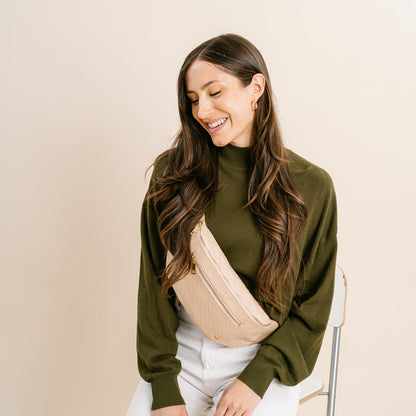 A woman with long brown hair in an olive green sweater and white pants smiles as she looks down, wearing The Darling Effect’s Woven Oat Trendy Luxe Belt Bag slung across her chest.