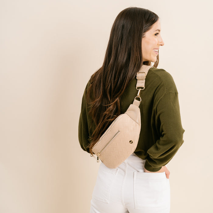 A woman with long brown hair, in a dark green sweater and white pants, stands sideways smiling. She wears The Darling Effect’s Woven Oat Trendy Luxe Belt Bag slung across her back. The background is plain off-white.