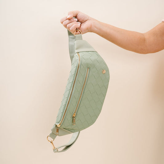 A person holds The Darling Effect's Woven Sage Trendy Luxe Belt Bag with gold zippers and hardware against a light beige background, highlighting this chic vegan leather hands-free accessory.