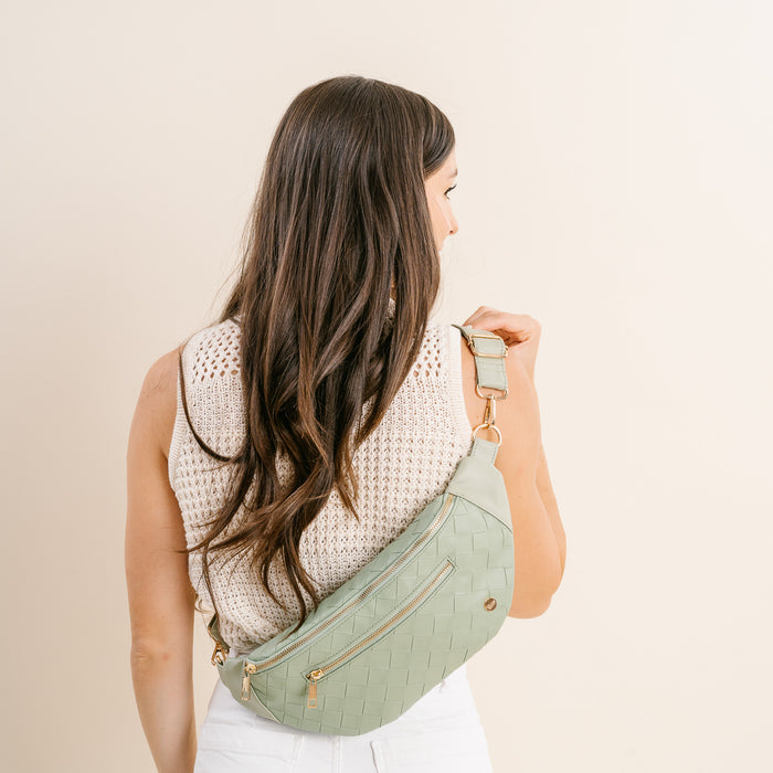 A woman with long brown hair, dressed in a sleeveless knit top and white pants, carries The Darling Effect’s Woven Sage Trendy Luxe Belt Bag—a stylish vegan leather crossbody—over her shoulder as she shows her back.