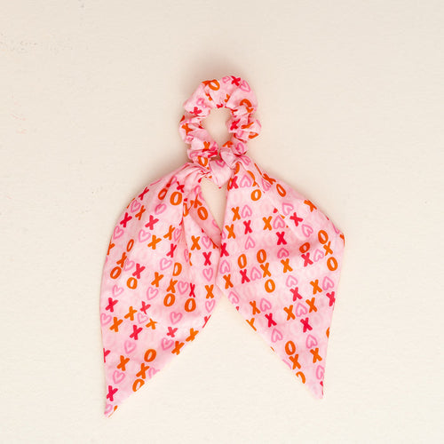 The XOXO Hair Scarf by the-darling-effect is a pink scrunchie with two scarf-like tails, displaying an XOXO pattern in pink, red, and orange on a plain off-white background.