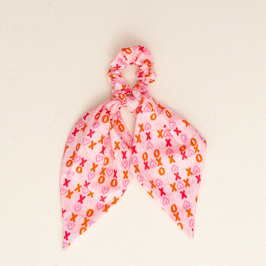 The XOXO Hair Scarf by the-darling-effect is a pink scrunchie with two scarf-like tails, displaying an XOXO pattern in pink, red, and orange on a plain off-white background.