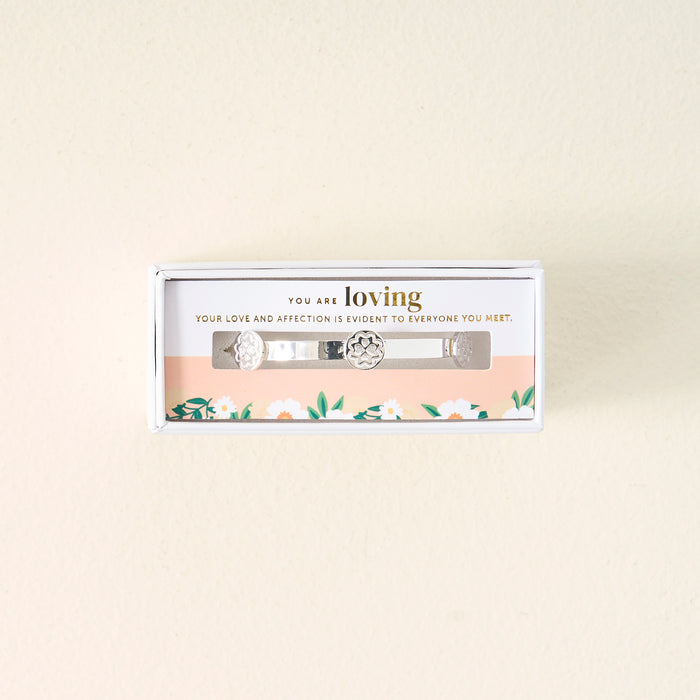 The Darling Effect’s You Are Loving Skinny Stacker Bracelet, a silver adjustable cuff with floral designs, arrives in a gift-ready box that reads “You are loving,” with flowers decorating the inside.