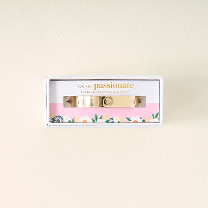 The Darling Effect’s You Are Passionate Cuff Bracelet features gold charms and comes boxed with floral-bordered packaging that reads, “You are passionate. Pursue what makes you happy,” set against a light background.