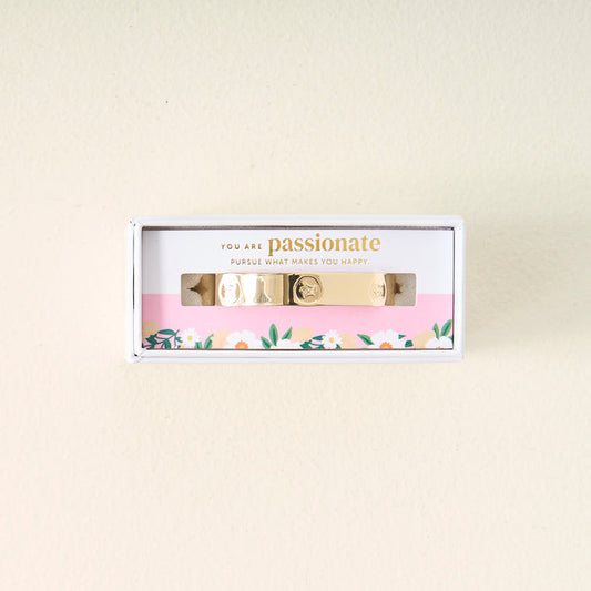 The Darling Effect’s You Are Passionate Cuff Bracelet features gold charms and comes boxed with floral-bordered packaging that reads, “You are passionate. Pursue what makes you happy,” set against a light background.