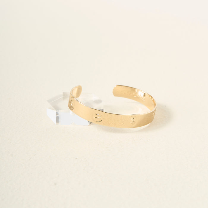 The Darling Effect’s You Are Passionate Cuff Bracelet, featuring engraved circular motifs, sits on a light surface next to a clear acrylic block, highlighting the timeless style of this adjustable accessory.