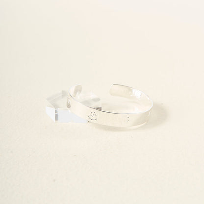 The Darling Effect’s "You Are Passionate" cuff bracelet in sterling silver, detailed with smiley face engravings, is displayed on a clear faceted acrylic block set against a light neutral background.