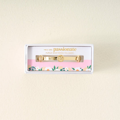 The Darling Effect’s You Are Passionate Skinny Stacker Bracelet features a 14k gold plated adjustable cuff with star and heart charms, presented in a floral-accented gift box. The lid reads: You are passionate. Pursue what makes you happy.