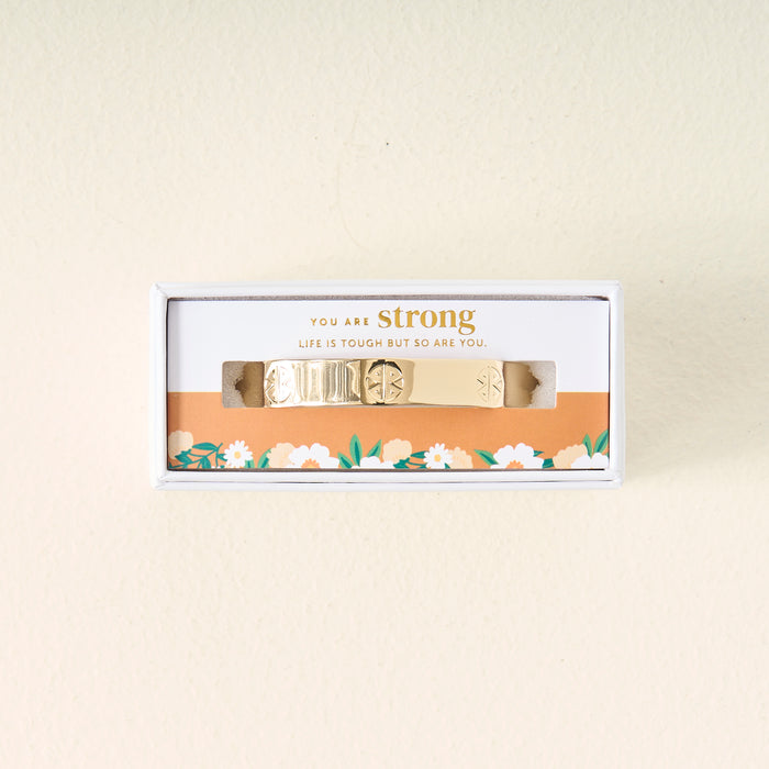 The Darling Effect's You Are Strong Cuff Bracelet features gold with unique patterns, presented in a white box with an orange floral design and the message "You are strong. Life is tough but so are you." Adjustable for elegant style.