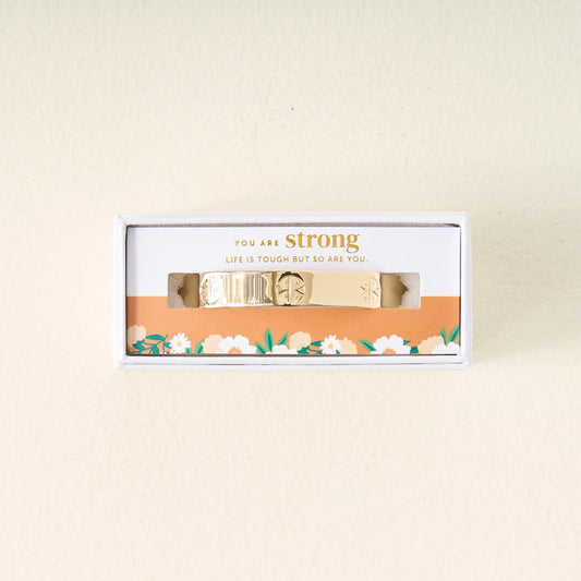 The Darling Effect's You Are Strong Cuff Bracelet features gold with unique patterns, presented in a white box with an orange floral design and the message "You are strong. Life is tough but so are you." Adjustable for elegant style.