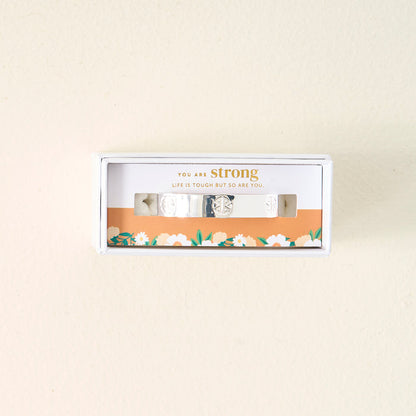 The Darling Effect's You Are Strong Cuff Bracelet, made of sterling silver, comes in a white floral box with “You are strong. Life is tough but so are you.” above the adjustable bracelet, set on a light beige background.