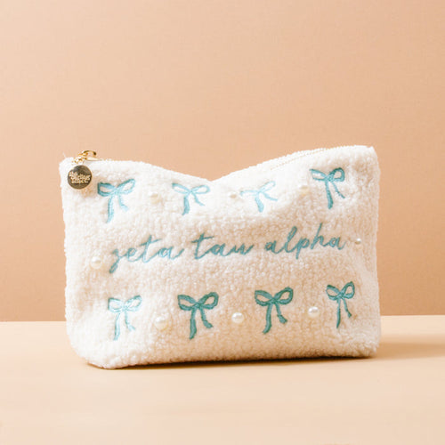 The Darling Effect's Zeta Tau Alpha Bows & Pearls Teddy Pouch features white sherpa with blue embroidered bows, pearls, and "zeta tau alpha" in blue—making it a perfect sorority gift.