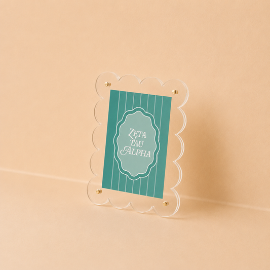 The Darling Effect’s Zeta Tau Alpha Mini Acrylic Picture Frame Magnet features scalloped edges and a teal card with white text, styled like a mini frame, and is shown propped against a neutral beige background.