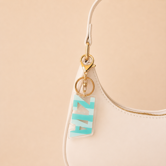 A white handbag with a gold clasp is shown against a beige background, featuring The Darling Effect’s Zeta Tau Alpha Stripes Acrylic Keychain with blue and white letters hanging from the handle.