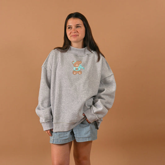 A woman with long brown hair wears The Darling Effect's Zeta Tau Alpha Teddy Bear Mockneck Sweatshirt in L/XL and light denim shorts, standing against a plain tan background.