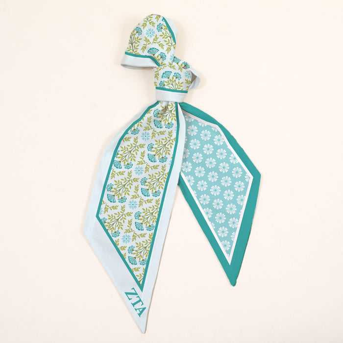 The Darling Effect's Zeta Tau Alpha Twirl & Tie Scarf Accessory features a green, blue, and white floral pattern with a geometric border and ZTA letters in one corner, all set on a light beige background.