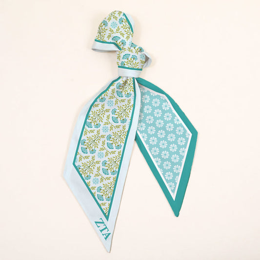 The Darling Effect's Zeta Tau Alpha Twirl & Tie Scarf Accessory features a green, blue, and white floral pattern with a geometric border and ZTA letters in one corner, all set on a light beige background.
